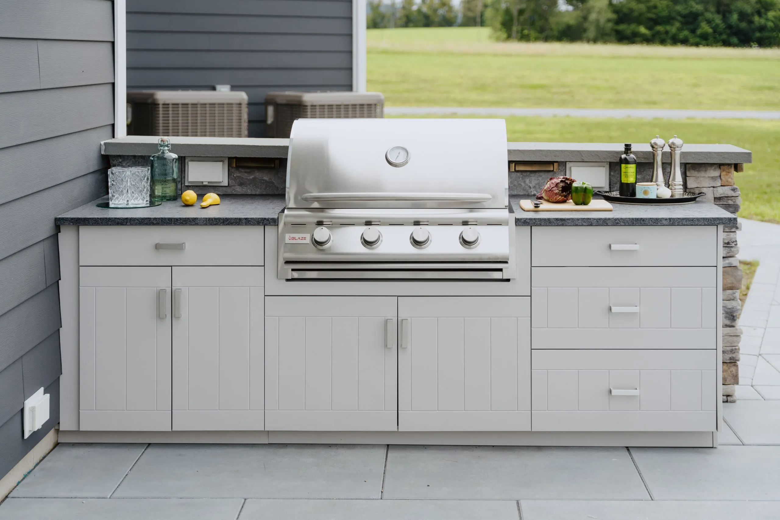 Open Air Cabinetry August Outdoor kitchen with light gray cabinetry, stainless steel grill, and dark stone countertops in a residential backyard setting