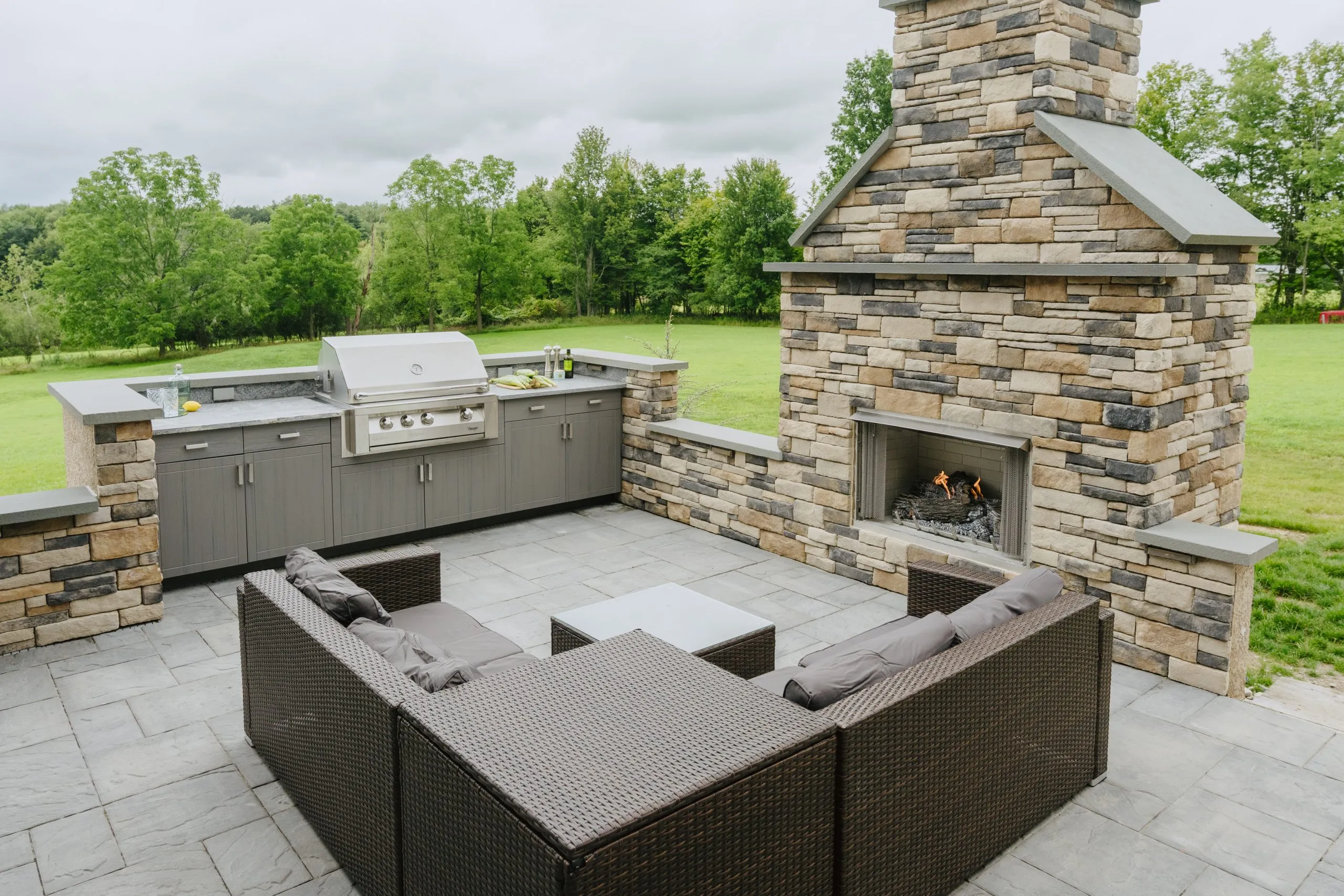 Open Air Cabinetry August Outdoor kitchen and fireplace with patio seating in landscaped yard