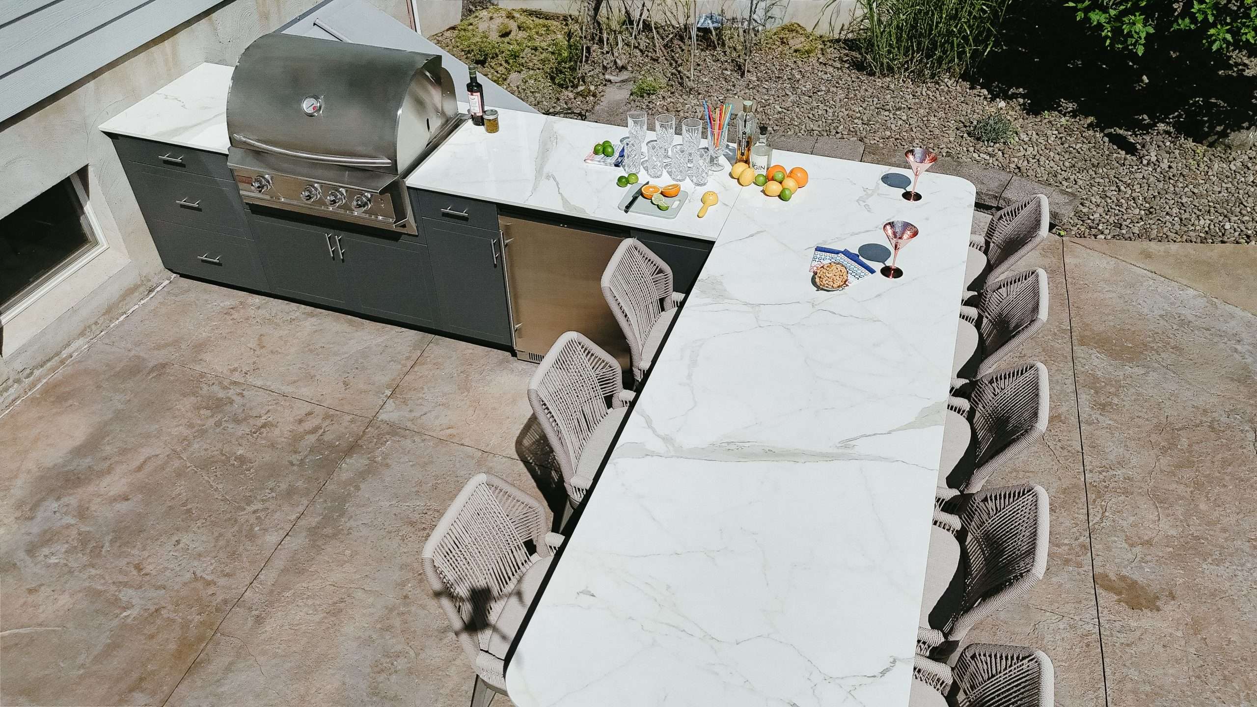 aQLNUVaO Outdoor kitchen and dining setup featuring dark cabinetry with stainless steel grill and an extended white marble countertop with woven chairs